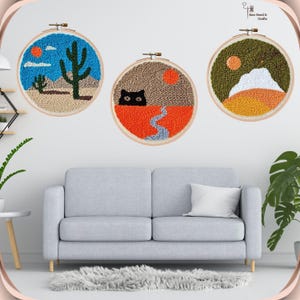 May include: Three embroidery hoop wall art pieces with desert scenes. The first hoop features a cactus and a blue sky. The second hoop features a black cat and a river. The third hoop features a mountain and a sunset.
