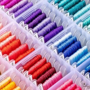 May include: A clear plastic organiser filled with spools of colourful embroidery floss. The floss is arranged by colour, including shades of pink, red, orange, yellow, purple, blue, and green. Each spool is labelled with a number. The image is well-lit and shows the variety of colours.