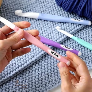 Puede incluir: Una colección de ganchillos de colores y tamaños variados. La imagen destaca un ganchillo rosa de 10,0 mm, con otros en morado, azul y verde. Los ganchillos están sobre una tela de ganchillo azul claro texturizada, para manualidades.