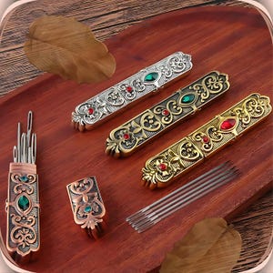 May include: Four ornate metal needle cases with jeweled accents and a set of needles. Each case features a different color scheme with green, red, and gold accents. The cases are arranged on a wooden surface.