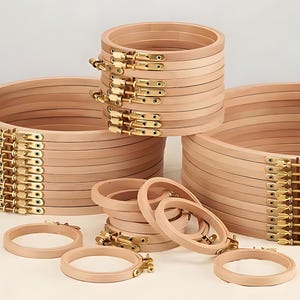 May include: A collection of wooden embroidery hoops in various sizes. The hoops are made of light-colored wood and feature brass-colored hardware. Several stacks of hoops are visible, along with individual hoops, suggesting a craft supply theme.