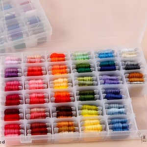 May include: A clear plastic storage box filled with colourful embroidery floss. The floss is arranged in a grid of small compartments, showcasing a spectrum of colours from pinks and reds to blues and greens. The text "Note: Floss Not Included" is visible.