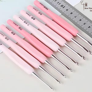 May include: A collection of pink crochet hooks with silver metal tips, arranged diagonally. Each hook has a size marking in millimetres. A metal ruler is in the background. The logo "Sew Need It Crafts" is in the upper right corner.