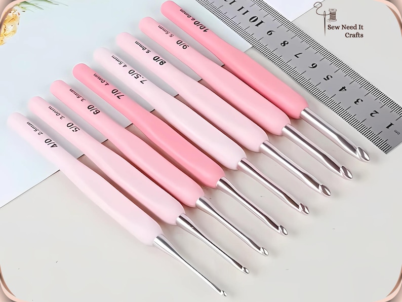 May include: A collection of pink crochet hooks with silver metal tips, arranged diagonally. Each hook has a size marking in millimetres. A metal ruler is in the background. The logo "Sew Need It Crafts" is in the upper right corner.