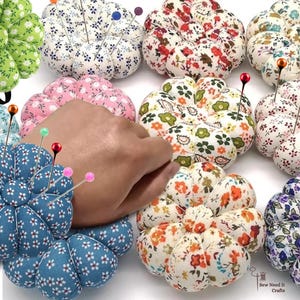 Op de afbeelding: Een collectie ronde, met stof beklede speldenkussens met diverse bloemmotieven in kleuren zoals groen, blauw, roze, oranje en paars. Elk kussen heeft een gesegmenteerde, bloemachtige vorm met ingestoken spelden. De tekst luidt Sew Need It Crafts.