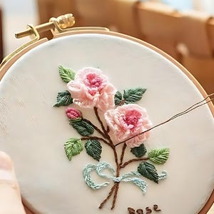 May include: Embroidered floral design on a white fabric stretched within a wooden hoop. The design features pink roses, green leaves, and a light blue bow. The word "rose" is stitched at the bottom. A hand is holding the hoop.