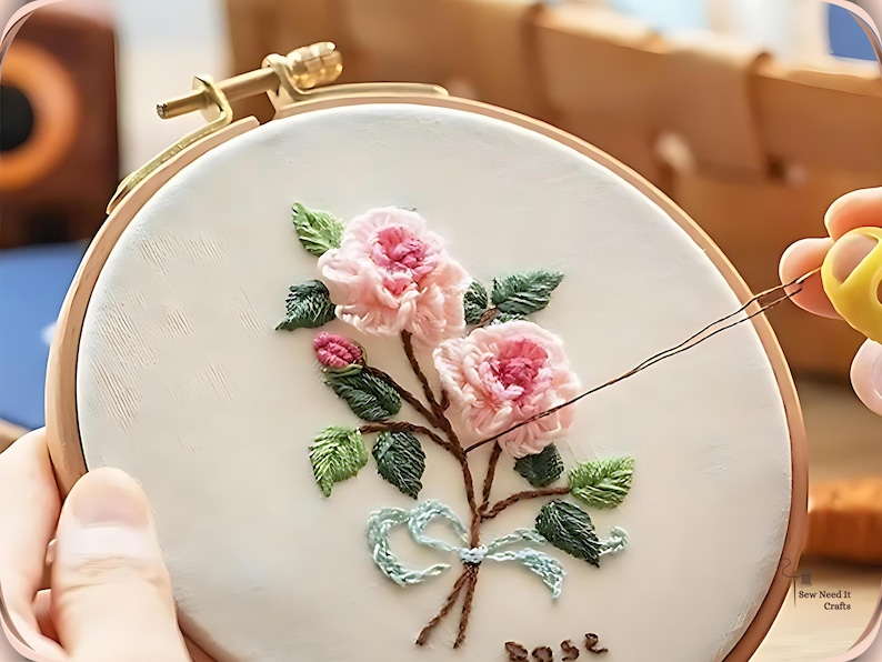May include: Embroidered floral design on a white fabric stretched within a wooden hoop. The design features pink roses, green leaves, and a light blue bow. The word "rose" is stitched at the bottom. A hand is holding the hoop.