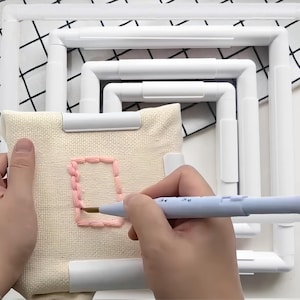 Könnte beinhalten: Ein Set aus ineinander verschachtelten, quadratischen Stickrahmen aus weißem Kunststoff. Ein Stück beiger Stoff ist im kleinsten Rahmen gespannt und zeigt eine mit hellrosa Garn gestickte rechteckige Form, gehalten von einem hellblauen Punch-Needle-Werkzeug.