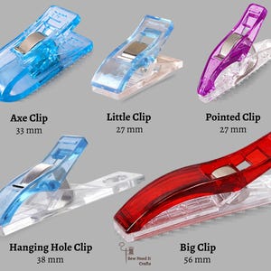 Op de afbeelding: Vijf transparante plastic naaiclips in verschillende kleuren: blauw, paars en rood. Elke clip heeft een zilveren metalen veer en is gelabeld met een naam en grootte in millimeters. De clips variëren in grootte van 27 mm tot 56 mm.