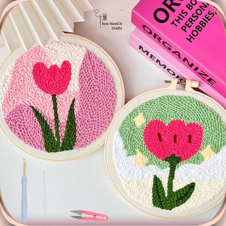 May include: Two embroidery hoops with pink and green yarn depicting a flower with a pink center and green leaves. The hoops are on a white surface with a pink book and embroidery tools.