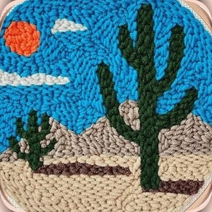 May include: A close-up of a needlepoint embroidery depicting a desert scene with a large green cactus, a smaller cactus, and a bright orange sun in a blue sky with white clouds.