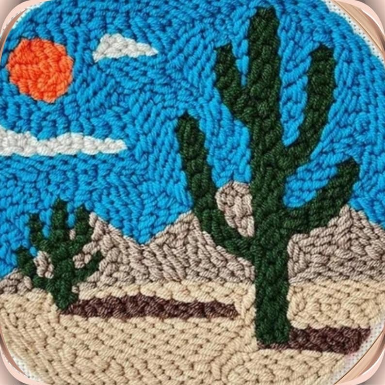 May include: A close-up of a needlepoint embroidery depicting a desert scene with a large green cactus, a smaller cactus, and a bright orange sun in a blue sky with white clouds.