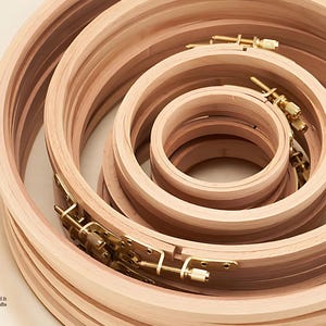 May include: A collection of wooden embroidery hoops in various sizes. The hoops are made of light-colored wood and feature brass hardware for tightening the fabric. The hoops are arranged in a nested spiral, showcasing their different diameters.