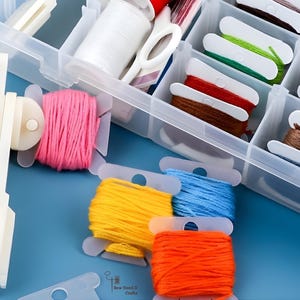 May include: A clear plastic craft organiser filled with spools of colourful embroidery floss in shades of pink, yellow, blue, and orange. A white thread winder and scissors are also visible.