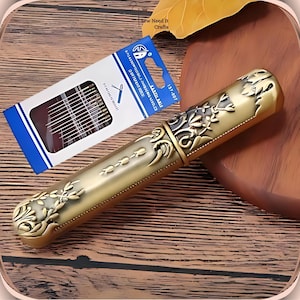 Op de afbeelding: Een gouden metalen naaldenkistje met een sierlijk bloemmotief. Het kistje is gesloten en heeft een scharnierend deksel. Het kistje ligt op een houten oppervlak. Het kistje is 15,7 cm lang.