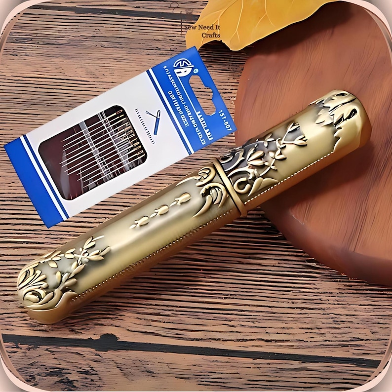 Op de afbeelding: Een gouden metalen naaldenkistje met een sierlijk bloemmotief. Het kistje is gesloten en heeft een scharnierend deksel. Het kistje ligt op een houten oppervlak. Het kistje is 15,7 cm lang.