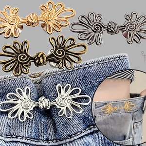May include: Four metal clothing fasteners in gold, silver, gunmetal, and antique bronze finishes. Each fastener features a symmetrical, open-work floral design with a central knot detail. One fastener is shown attached to blue denim jeans.