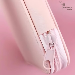 May include: Close-up of a pale pink zippered pouch. The zip and pull tab are the same colour as the pouch. The pouch is made of a smooth material. The words "Sew Need It Crafts" are in the upper right corner.