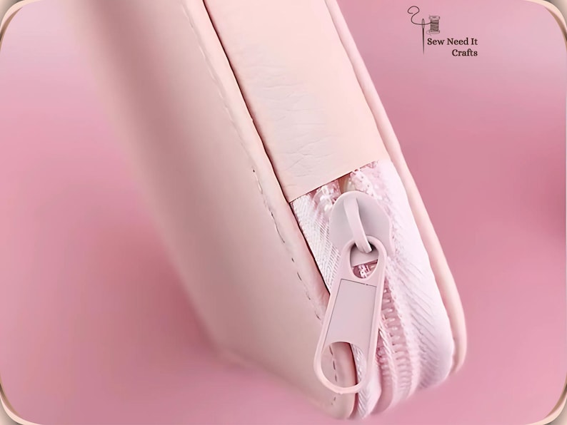 May include: Close-up of a pale pink zippered pouch. The zip and pull tab are the same colour as the pouch. The pouch is made of a smooth material. The words "Sew Need It Crafts" are in the upper right corner.