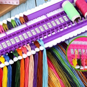 Può includere: Un organizer per fili viola con bordi smerlati bianchi, contenente fili da ricamo di vari colori. I fili sono disposti per colore, con fessure numerate per ogni filo. Include anche rocchetti di filo e un metro a nastro.