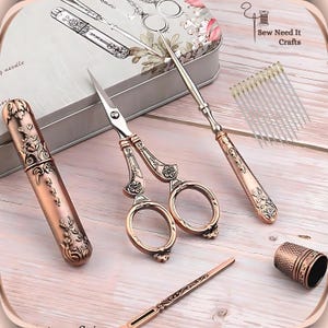 Può includere: Un set di strumenti da cucito in stile vintage con finitura in rame. Il set include un paio di forbici, un infila-ago, un ditale e una custodia per aghi. Gli strumenti sono disposti su una superficie in legno. Sew Need It Crafts.