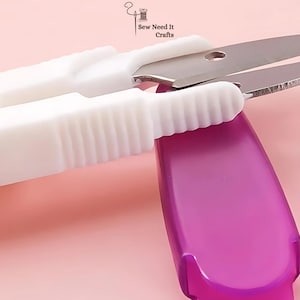 May include: Close-up of white and silver thread snips with a purple handle. The scissors are open, ready to cut thread. The background is a soft pink.