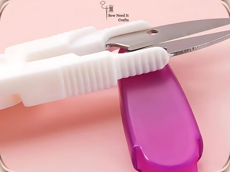 May include: Close-up of white and silver thread snips with a purple handle. The scissors are open, ready to cut thread. The background is a soft pink.
