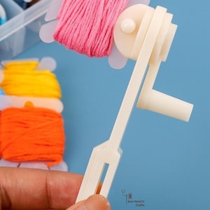 May include: A white thread winder tool is held in front of a blue background. Spools of colourful embroidery floss in pink, yellow, orange, and blue are visible. The tool is designed to wind thread onto bobbins.