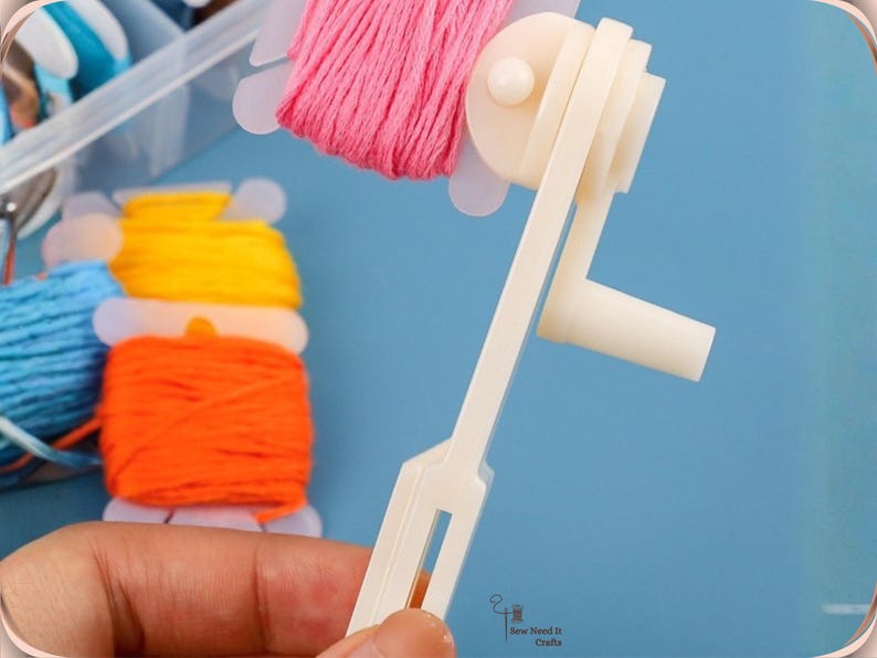 May include: A white thread winder tool is held in front of a blue background. Spools of colourful embroidery floss in pink, yellow, orange, and blue are visible. The tool is designed to wind thread onto bobbins.