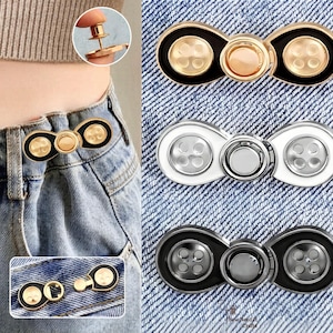Puede incluir: Un conjunto de tres extensores de botón de jean decorativos en dorado, plateado y negro. Cada extensor presenta un diseño de botón con un botón central y botones decorativos más pequeños. Los extensores se muestran sobre tela vaquera.