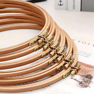 May include: A stack of wooden embroidery hoops with brass hardware. The hoops are light brown and arranged in a fan shape, with the brass fasteners visible. The background includes a printed page with the word "VILLAGE".