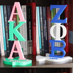 May include: Four different sorority letter plaques, each with a different color scheme. The plaques are made of plastic and feature the letters of the sorority in a 3D design. The plaques are displayed on a bookshelf with a variety of books behind them.