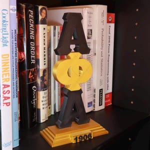 May include: A black and gold 3D letter sculpture of the Greek letters "Alpha Phi Alpha" with the year "1906" on a gold base. The sculpture is on a bookshelf with other books.