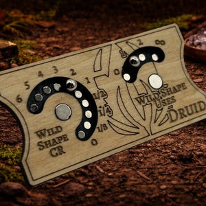 以下が含まれることがあります： 「Wild Shape Uses Druid」と「Wild Shape CR」の文字が入った木製のゲームボード。ボードには、磁石のピースとエッチングされたデザインが施された曲線トラックがあります。背景は森の床です。