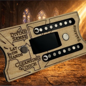 May include: A wooden Paladin device with engraved text reading "Divine Sense", "Lay on Hands", and "Cleansing Touch". It has a black screen, silver buttons, and rows of small silver dots. The device is set against a blurred background.