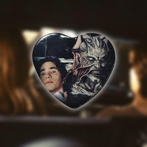 May include: Heart-shaped button featuring a horror movie scene. The button showcases a man and a monster in a dark setting. The button is black with a glossy finish.