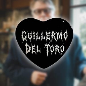 May include: A black heart-shaped object with the name "GUILLERMO DEL TORO" in white, stylized, gothic lettering. The object is glossy and appears to be a pin or a decorative item. The background is blurred.