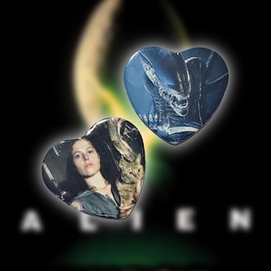 May include: Two heart-shaped Alien movie-themed pins. One pin features a portrait of Sigourney Weaver with a cat, the other depicts the Alien creature. The background is black with the word "ALIEN" in white.