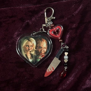 May include: A heart-shaped keychain featuring a printed image of two dolls, one with blonde hair and one with scarred skin. The keychain includes a silver lobster clasp, a red heart charm, a silver-toned knife charm with red paint, and beads.