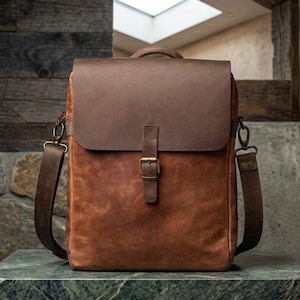 Può includere: Borsa messenger in pelle marrone con chiusura a patta e tracolla regolabile. La borsa presenta un manico superiore e una chiusura con fibbia. La pelle ha un aspetto invecchiato, con variazioni di colore. Circa 30 cm di larghezza.