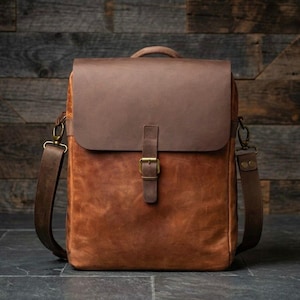 Può includere: Una borsa in pelle marrone con chiusura a patta e tracolla regolabile. La borsa presenta una maniglia superiore e una chiusura con fibbia sul davanti. La pelle ha un aspetto invecchiato e strutturato. La borsa è alta circa 35 cm e larga 30 cm.