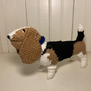 May include: A knitted plush toy dog in the shape of a basset hound. The dog is brown, black, and white with a blue collar.