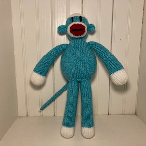 May include: A blue knitted monkey toy with a red mouth and white accents. The monkey has a long tail and is standing on a white surface.