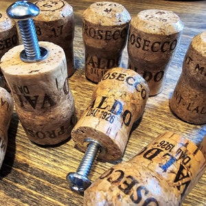 May include: A collection of used corks, some with metal screws inserted, arranged on a wooden surface. The corks have printed text, including "PROSECCO" and "VALDO DAL 1926".
