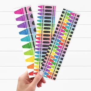 May include: Three colorful crayon-themed bookmarks. Each bookmark features a repeating pattern of crayons in various colors, including pink, purple, blue, green, yellow, orange, and red. The bookmarks are held by a hand against a white background.