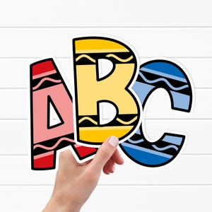 Op de afbeelding: Kleurrijke ABC-letters in een krijtstijl. De letters A, B en C worden door een hand vastgehouden, met andere letters zichtbaar op de achtergrond. De letters zijn roze, geel en blauw met zwarte golvende lijnen, die op kleurpotloden lijken.