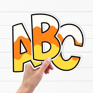 Op de afbeelding: ABC-letters met een candy corn-thema. De letters A, B en C worden door een hand vastgehouden. De letters zijn wit met oranje en gele golvende strepen, omlijnd in zwart. Extra letters zijn zichtbaar.