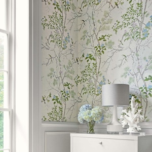 May include: A white wall with a floral wallpaper featuring green branches, blue flowers, and small birds. A white dresser with a gray lamp and a vase of blue flowers sits in front of the wall.