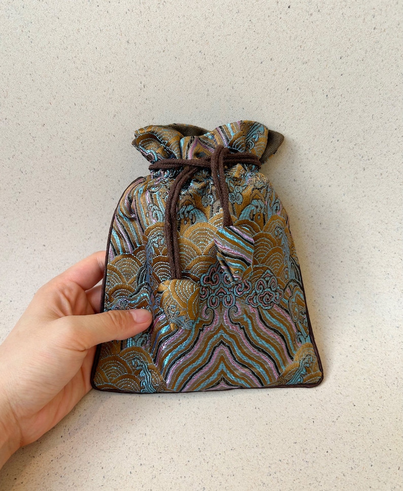 Puede incluir: Una peque&ntilde;a bolsa de cord&oacute;n hecha de tela marr&oacute;n con un dise&ntilde;o estampado azul y dorado. La bolsa tiene un cord&oacute;n marr&oacute;n y est&aacute; atada en la parte superior.