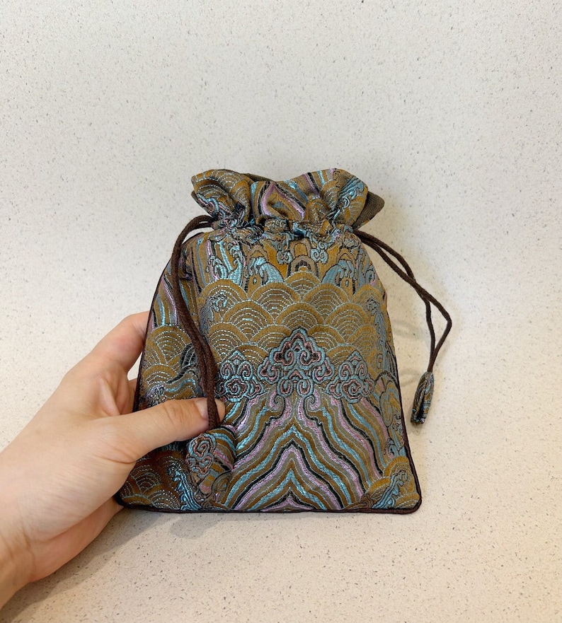 Puede incluir: Una peque&ntilde;a bolsa de cord&oacute;n con un cord&oacute;n marr&oacute;n y una tela estampada en azul, dorado y rosa. El patr&oacute;n presenta olas y nubes estilizadas.